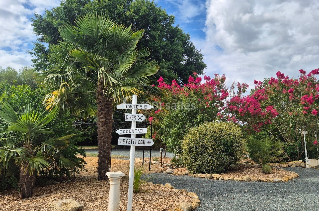 Panneau directionnel en bois dans un jardin arboré avec palmiers, fleurs roses et ciel partiellement nuageux.