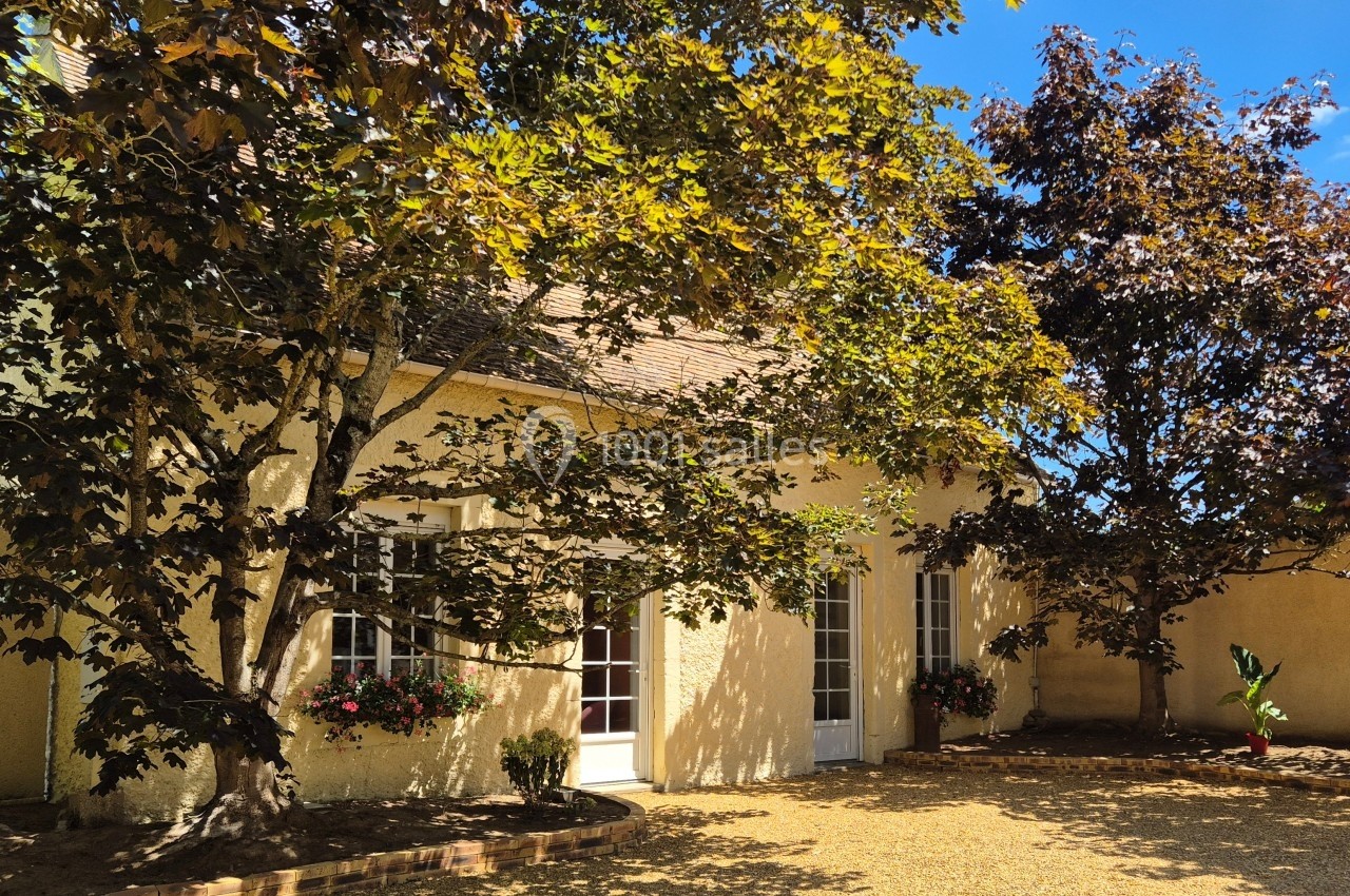 Façade d'une maison en pierre claire avec des arbres ombrageant une cour en gravier, par une journée ensoleillée.