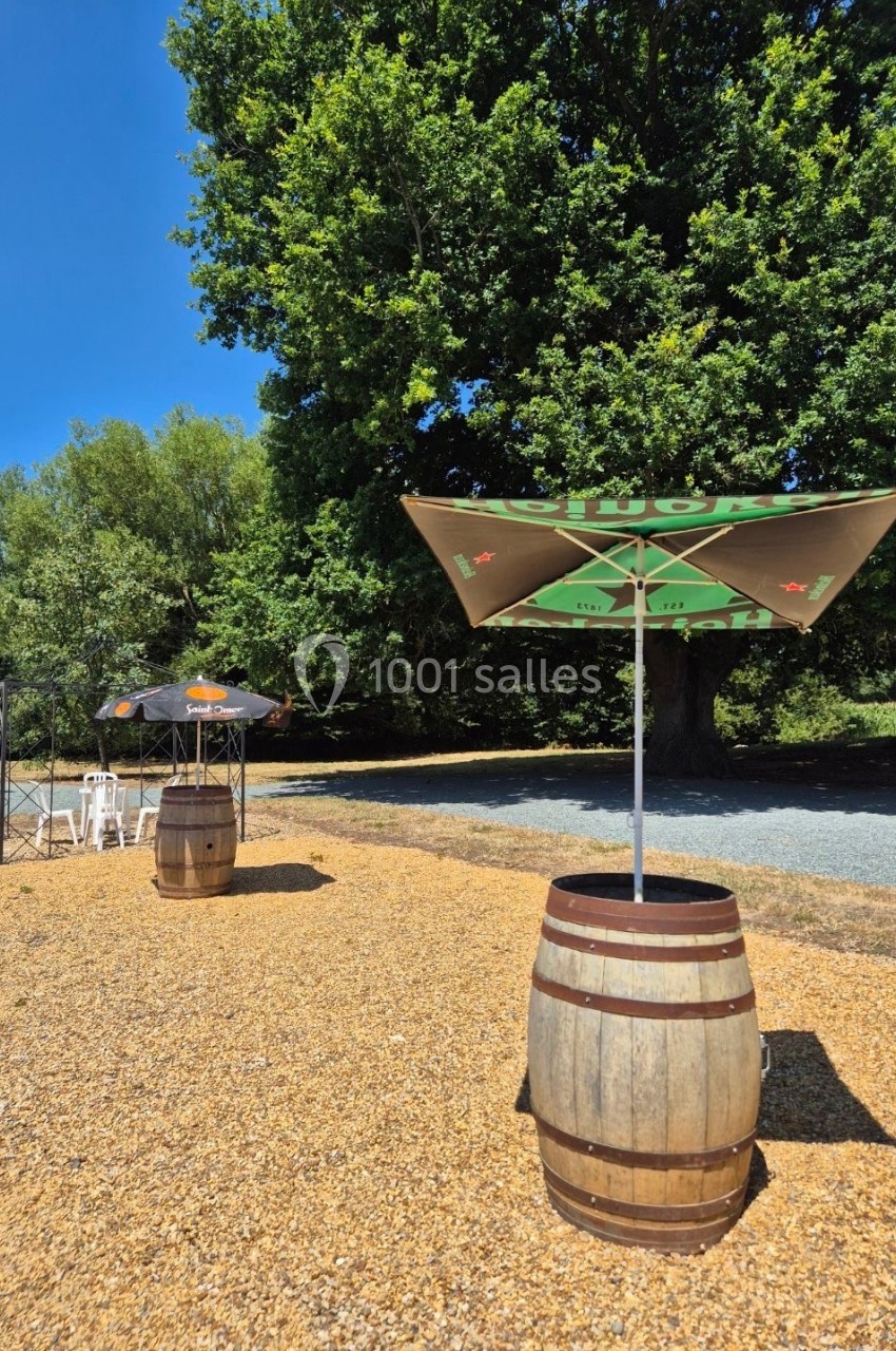 Deux tonneaux en bois avec parasols sur un sol gravillonné, près d'arbres sous un ciel bleu.
