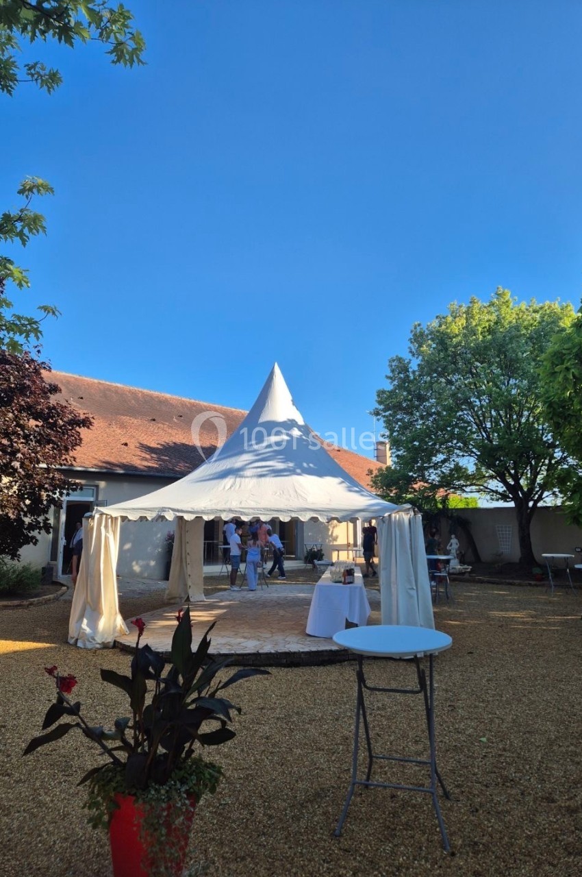 Tente blanche installée dans une cour extérieure ensoleillée, entourée d'arbres et de tables hautes.