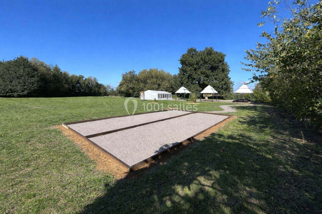Terrain de pétanque en gravier entouré d'une pelouse, avec des arbres et des structures blanches en arrière-plan.