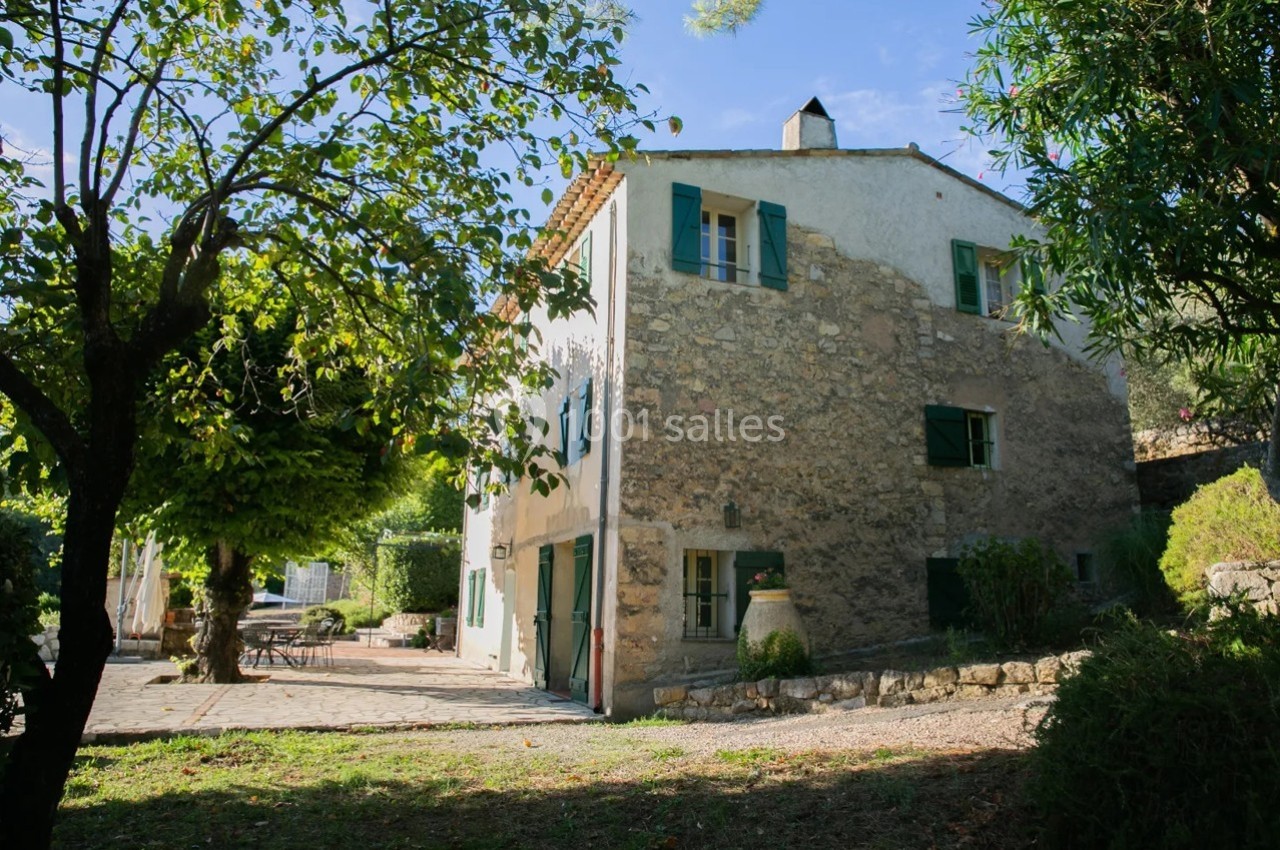 Maison en pierre avec volets verts, entourée de végétation et d'une terrasse ensoleillée.