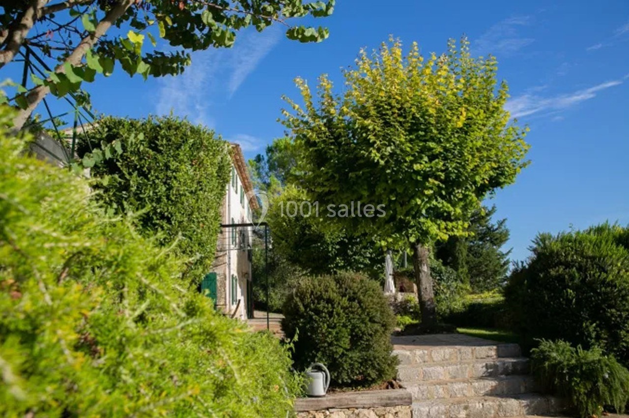Maison entourée de verdure avec un escalier en pierre menant à un jardin arboré sous un ciel bleu.
