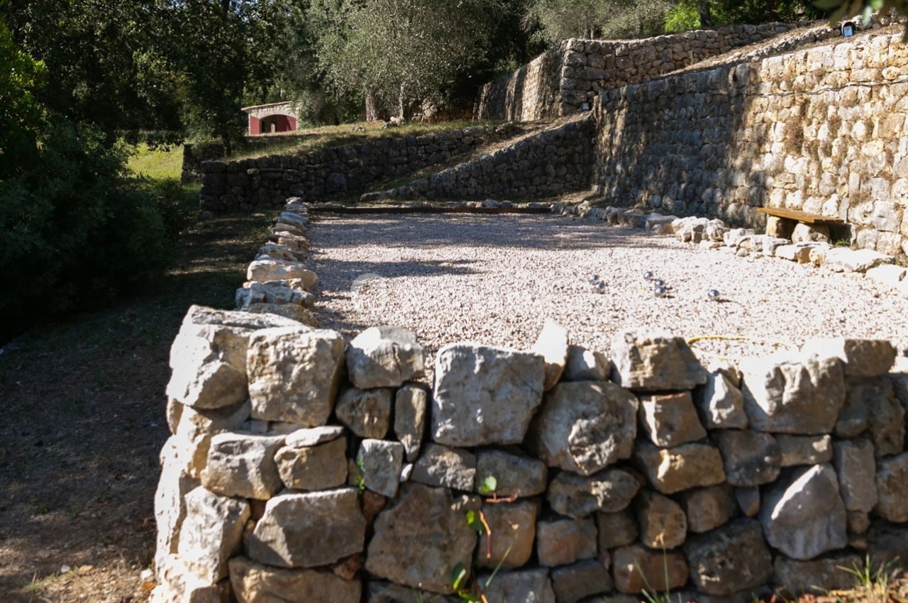 Mur en pierres sèches entourant une surface gravillonnée, avec des arbres et un petit bâtiment en arrière-plan.