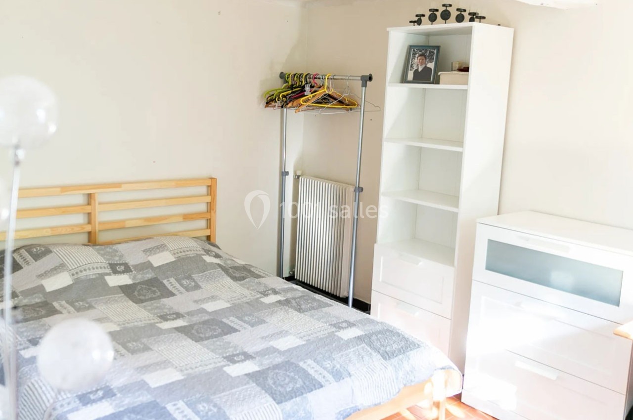 Chambre lumineuse avec lit double, étagère blanche, portant à vêtements et carrelage au sol.