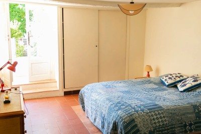 Chambre lumineuse avec lit double, couvre-lit bleu, lampe de chevet et porte-fenêtre donnant sur un jardin.