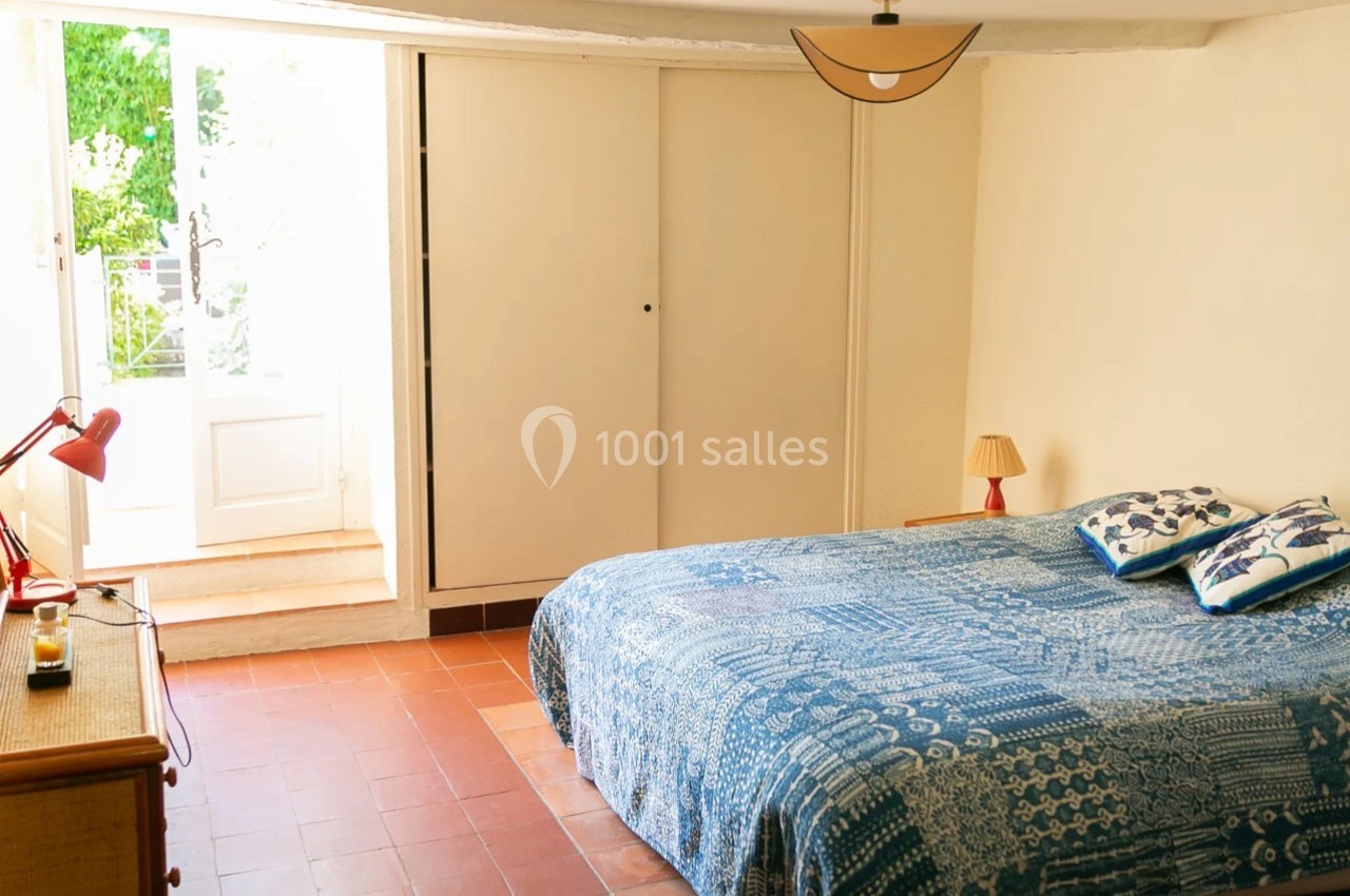 Chambre lumineuse avec lit double, couvre-lit bleu, lampe de chevet et porte-fenêtre donnant sur un jardin.