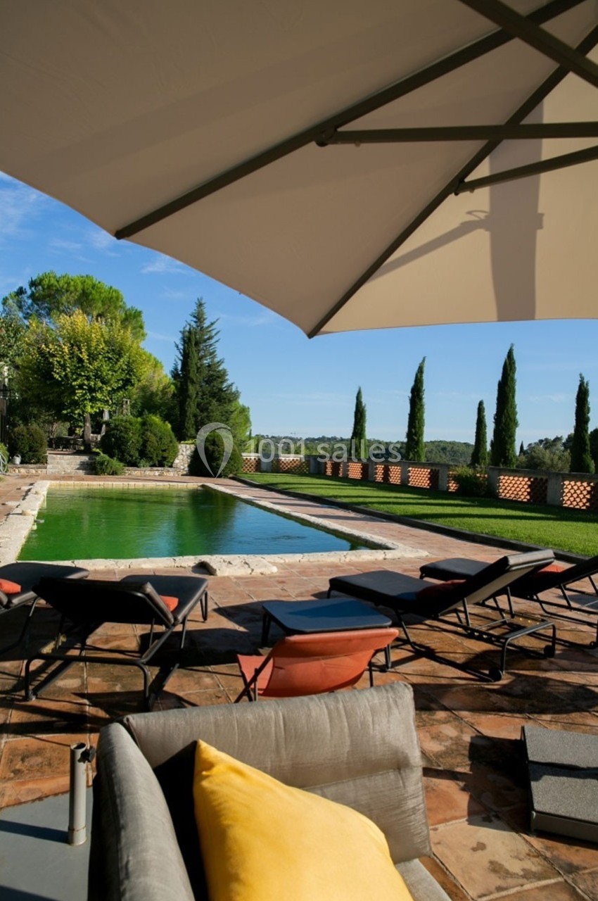 Piscine extérieure entourée de transats et d'arbres, partiellement ombragée par un grand parasol.