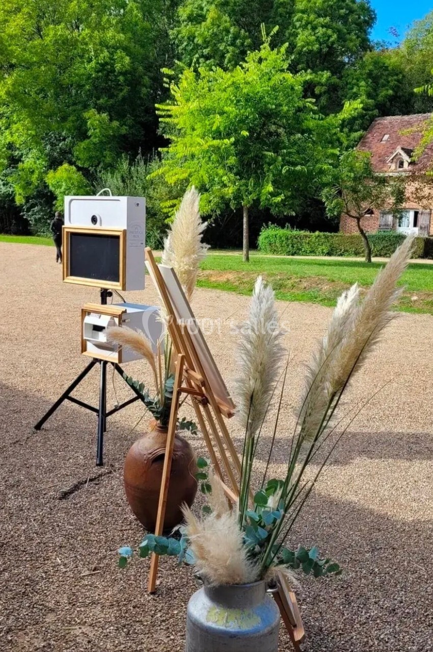 Photobooth installé en extérieur sur une allée gravillonnée, entouré de végétation et de décorations florales.