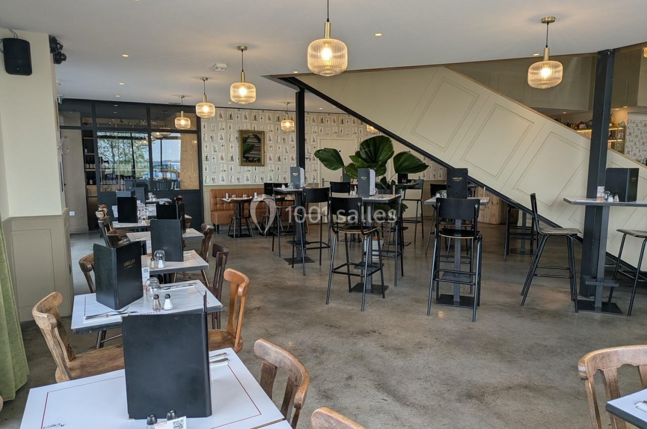 Salle de restaurant moderne avec tables dressées, chaises hautes, escalier central et décoration lumineuse.