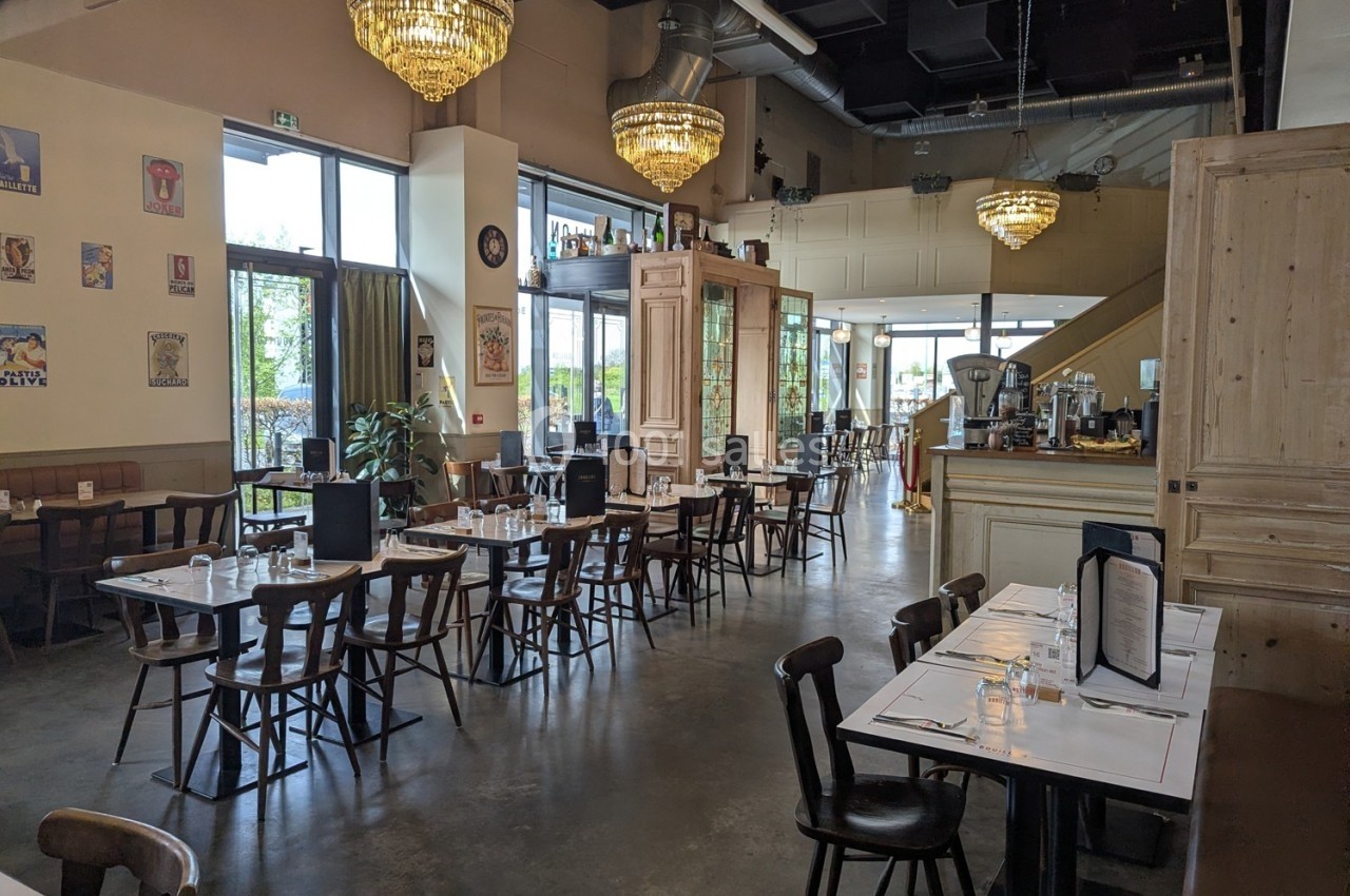 Salle de restaurant lumineuse avec tables dressées, chaises en bois, lustres suspendus et décorations murales.