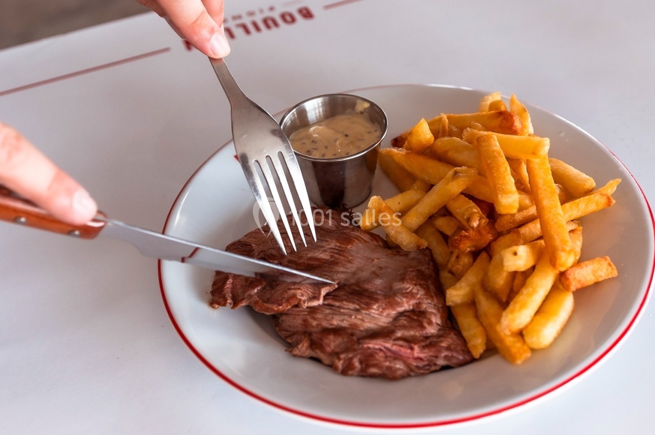 Assiette contenant un steak, des frites dorées et une sauce servie dans un ramequin, avec une personne découpant la viande.
