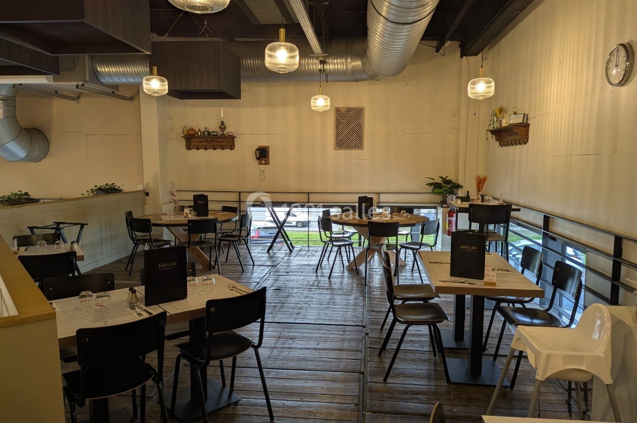 Salle de restaurant lumineuse avec tables dressées, chaises noires et grandes baies vitrées donnant sur l'extérieur.