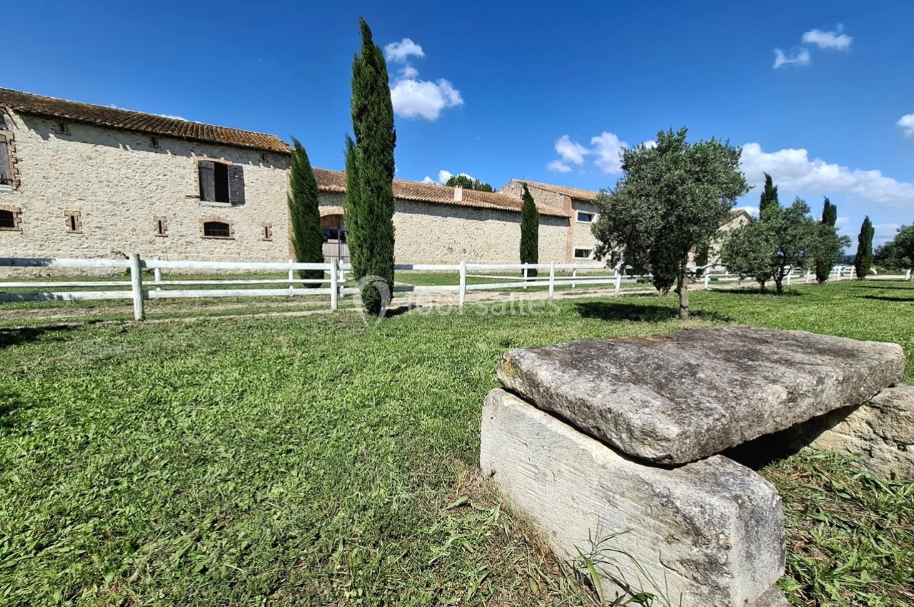 Banc en pierre dans un jardin verdoyant, avec en arrière-plan un bâtiment en pierre et des arbres sous un ciel bleu.