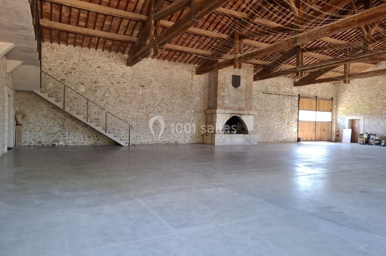 Intérieur d'une grande pièce avec murs en pierre, poutres apparentes, cheminée et sol en béton poli.