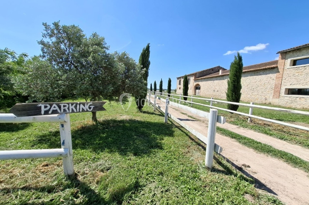 Chemin en gravier bordé de barrières blanches menant à un bâtiment en pierre, avec un panneau indiquant ’Parking’.