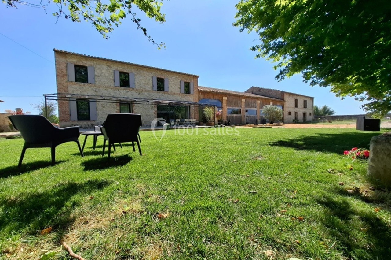 Grande maison en pierre avec jardin verdoyant, chaises noires sur la pelouse et arbres offrant de l'ombre.