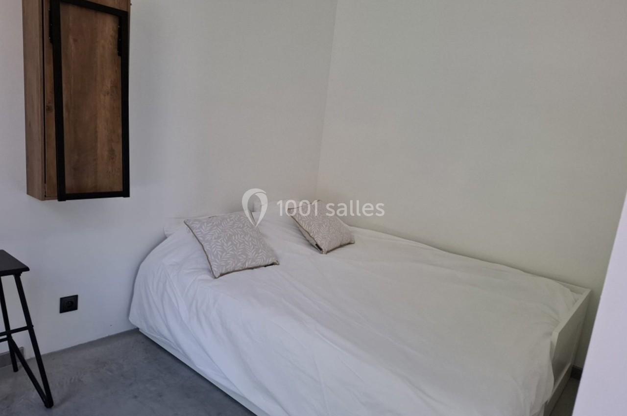Lit double avec draps blancs et deux coussins décoratifs dans une pièce aux murs clairs et sol en béton.