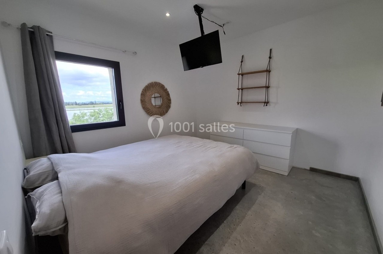 Chambre lumineuse avec lit double, commode blanche, étagères murales, fenêtre donnant sur un paysage et télévision suspendue.