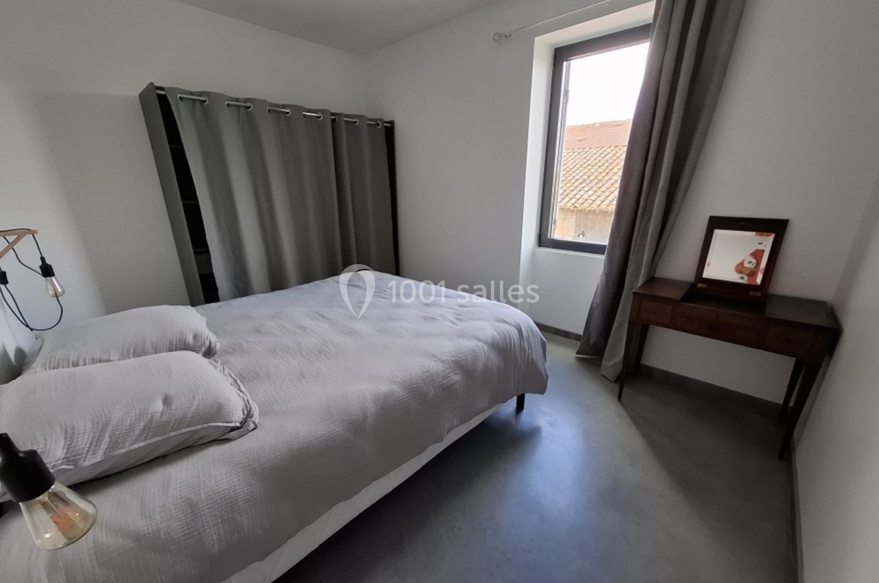 Chambre minimaliste avec lit double, rideaux gris, bureau en bois et fenêtre donnant sur un toit en tuiles.