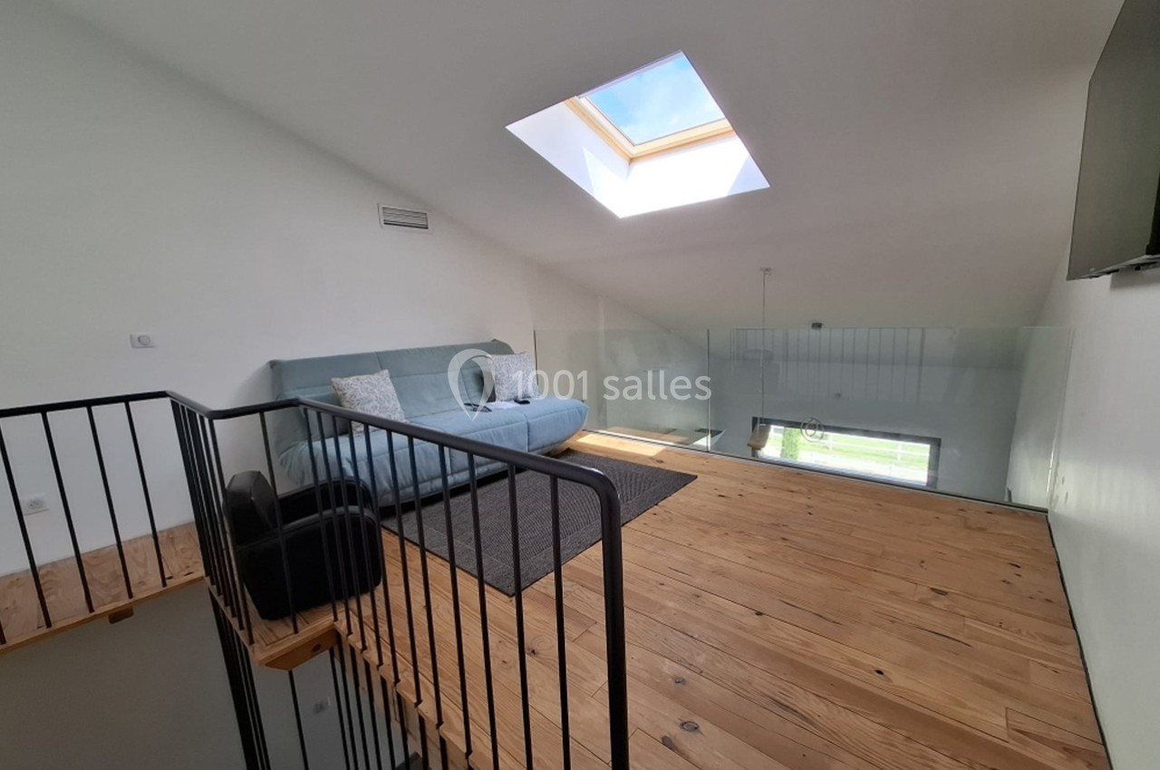 Mezzanine lumineuse avec parquet en bois, canapé, tapis gris et verrière donnant sur l’espace en dessous.