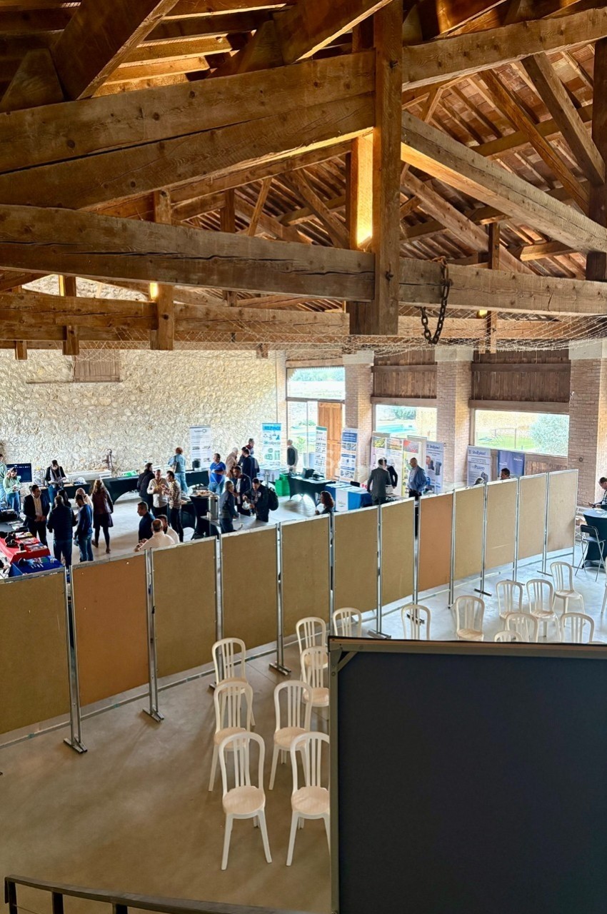 Vue d'une salle avec charpente en bois, des stands d'exposition, des visiteurs et des chaises blanches alignées.
