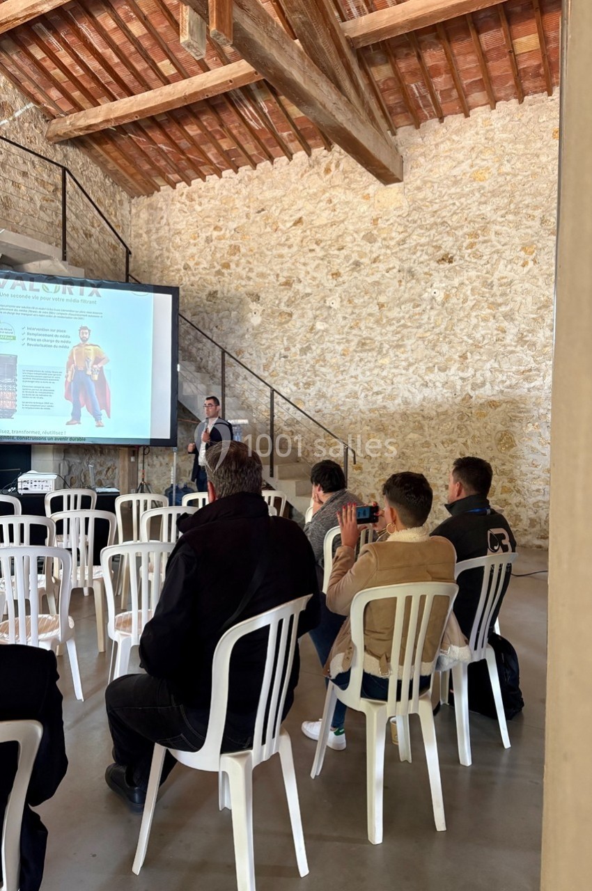 Un conférencier présente devant un petit public assis dans une salle en pierre avec un écran de projection.