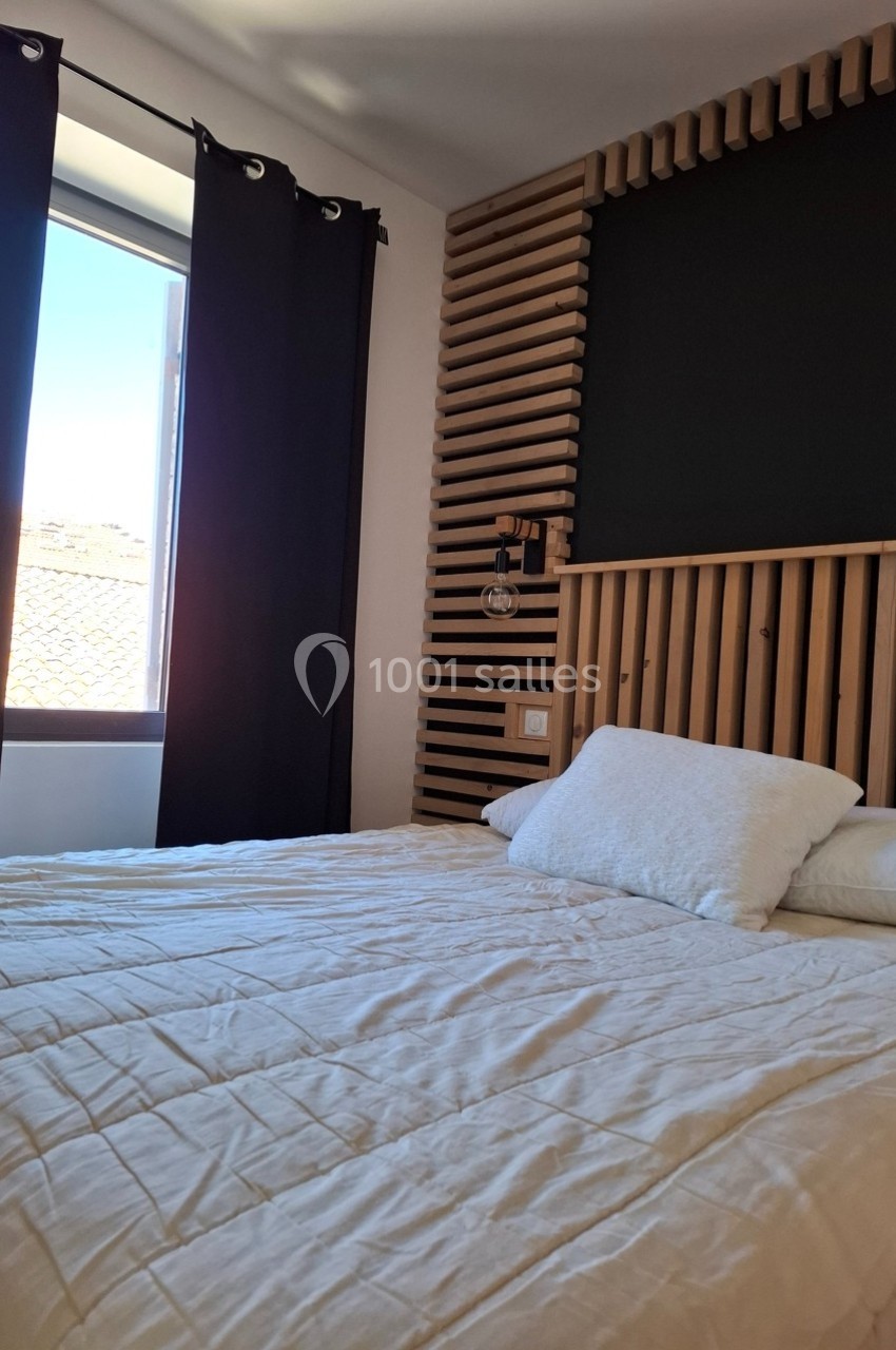Lit double avec couverture claire, tête de lit en bois, lampe murale et fenêtre avec rideaux noirs laissant entrer la…