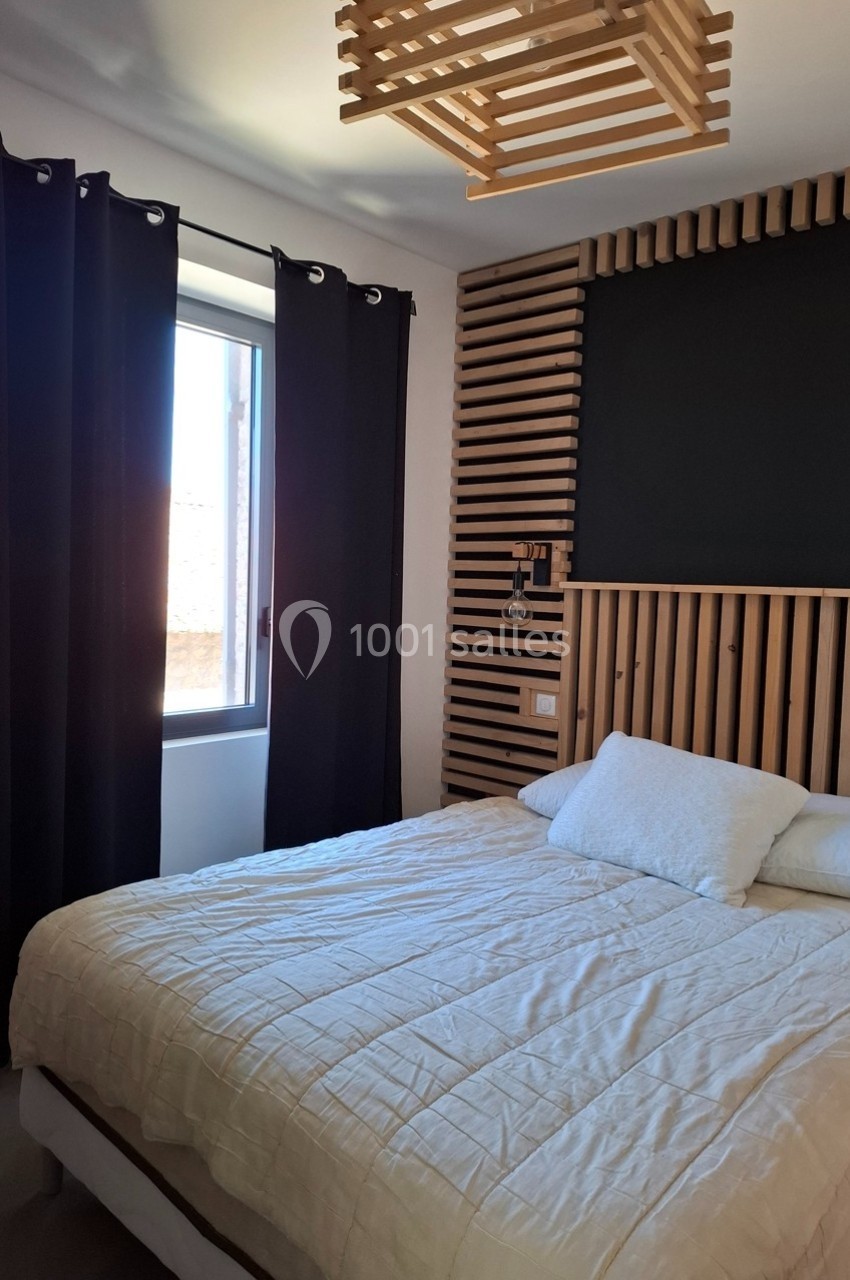 Chambre lumineuse avec lit double, tête de lit en bois, rideaux sombres et fenêtre laissant entrer la lumière naturelle.