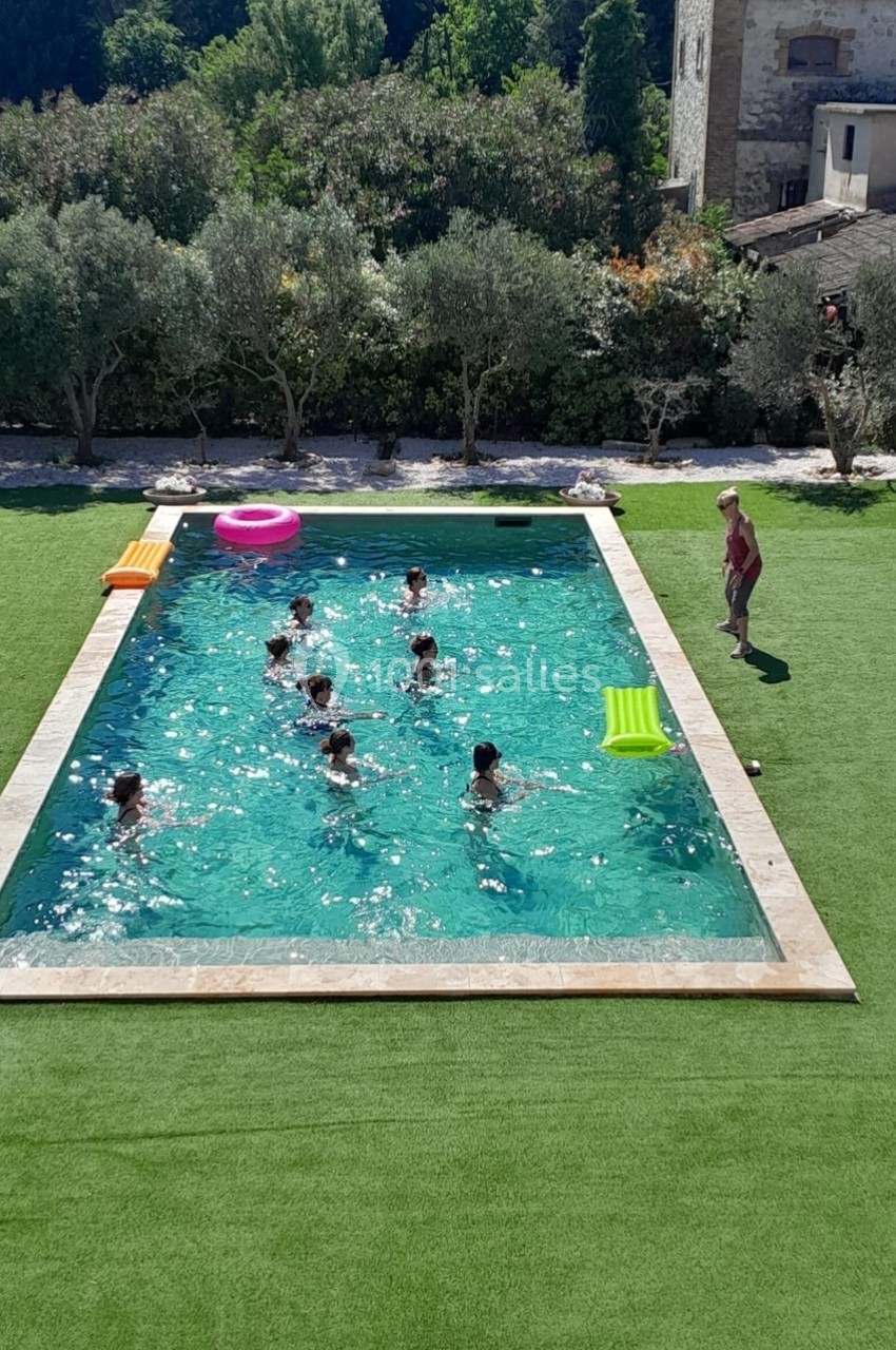 Vue aérienne d'une piscine extérieure entourée de pelouse, avec plusieurs personnes nageant et un homme debout au bord.