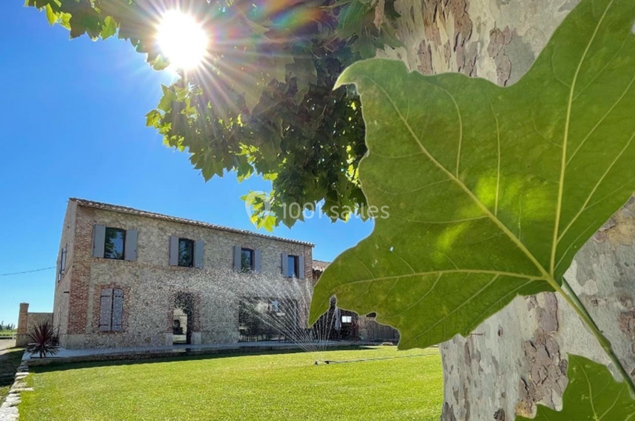 Vue d'une maison en pierre avec un jardin verdoyant, des arbres et un arrosage automatique sous un soleil éclatant.
