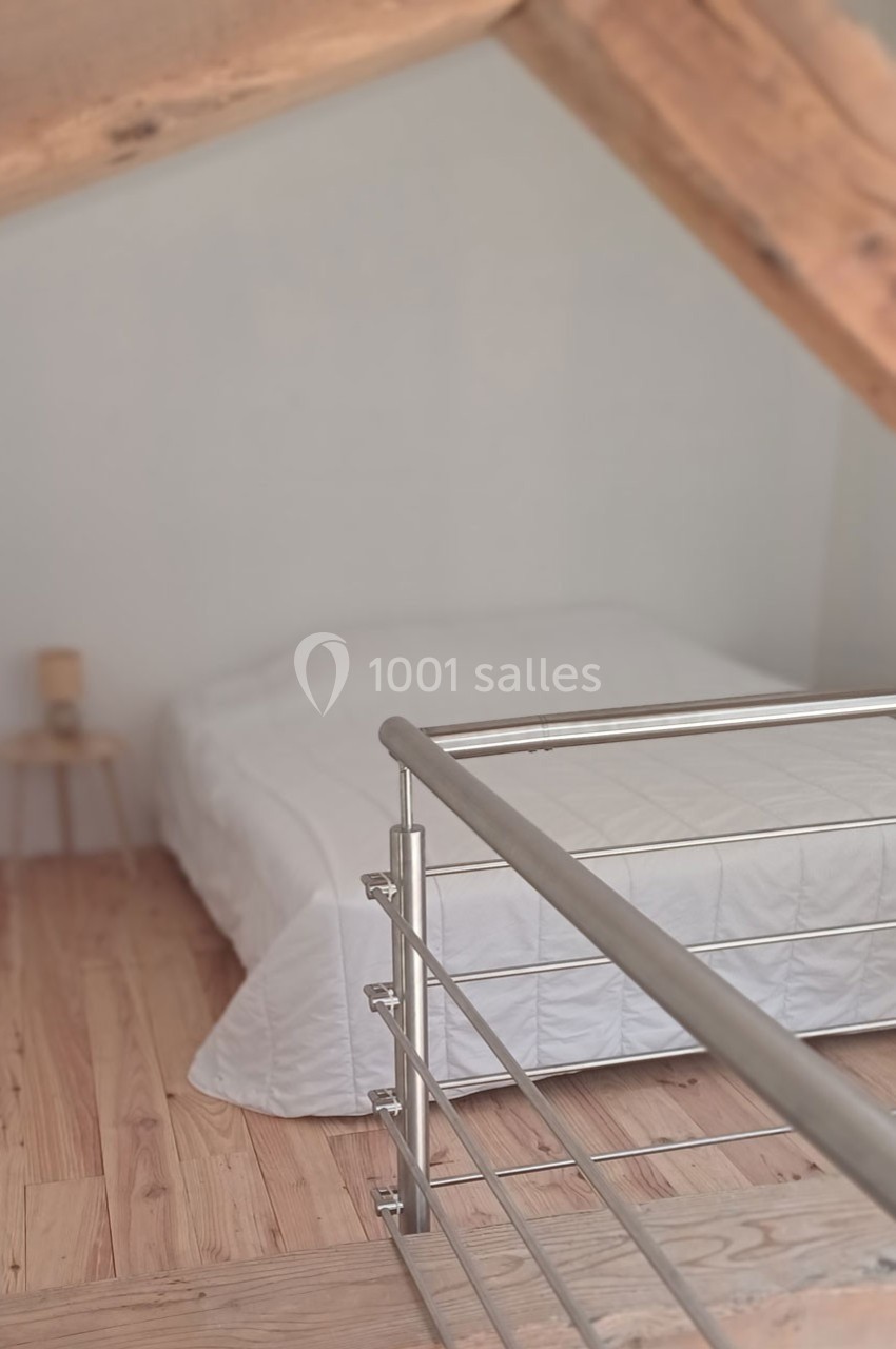 Lit double avec drap blanc dans une chambre mansardée au sol en bois clair, vue depuis une rambarde métallique.