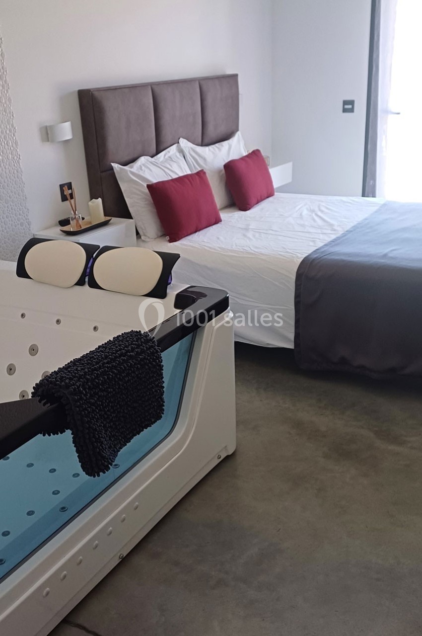 Chambre moderne avec lit double, coussins rouges, baignoire balnéo et lumière naturelle entrant par une baie vitrée.