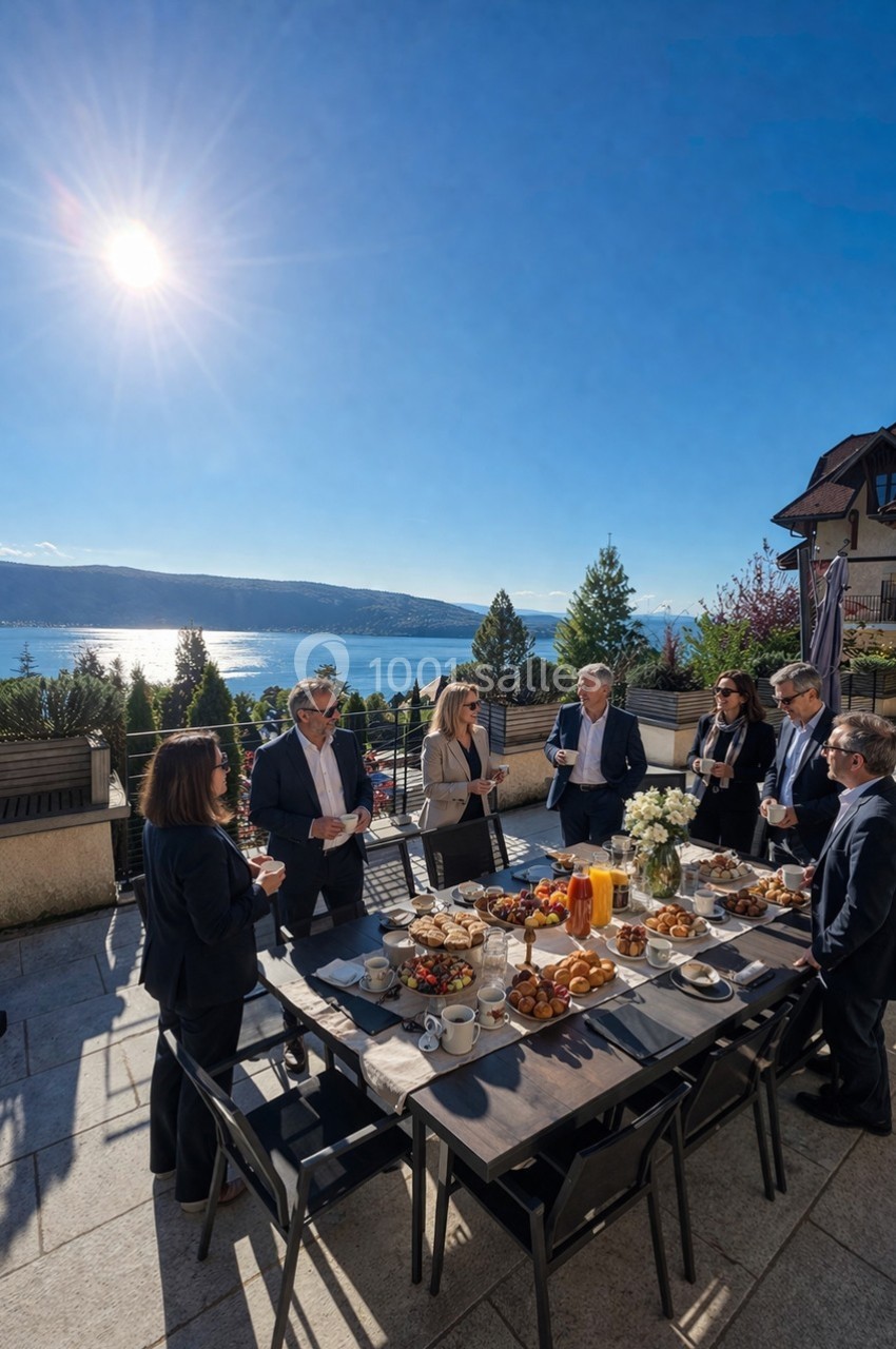 Groupe de personnes debout autour d'une table garnie de nourriture, en extérieur avec vue sur un lac et des montagnes.