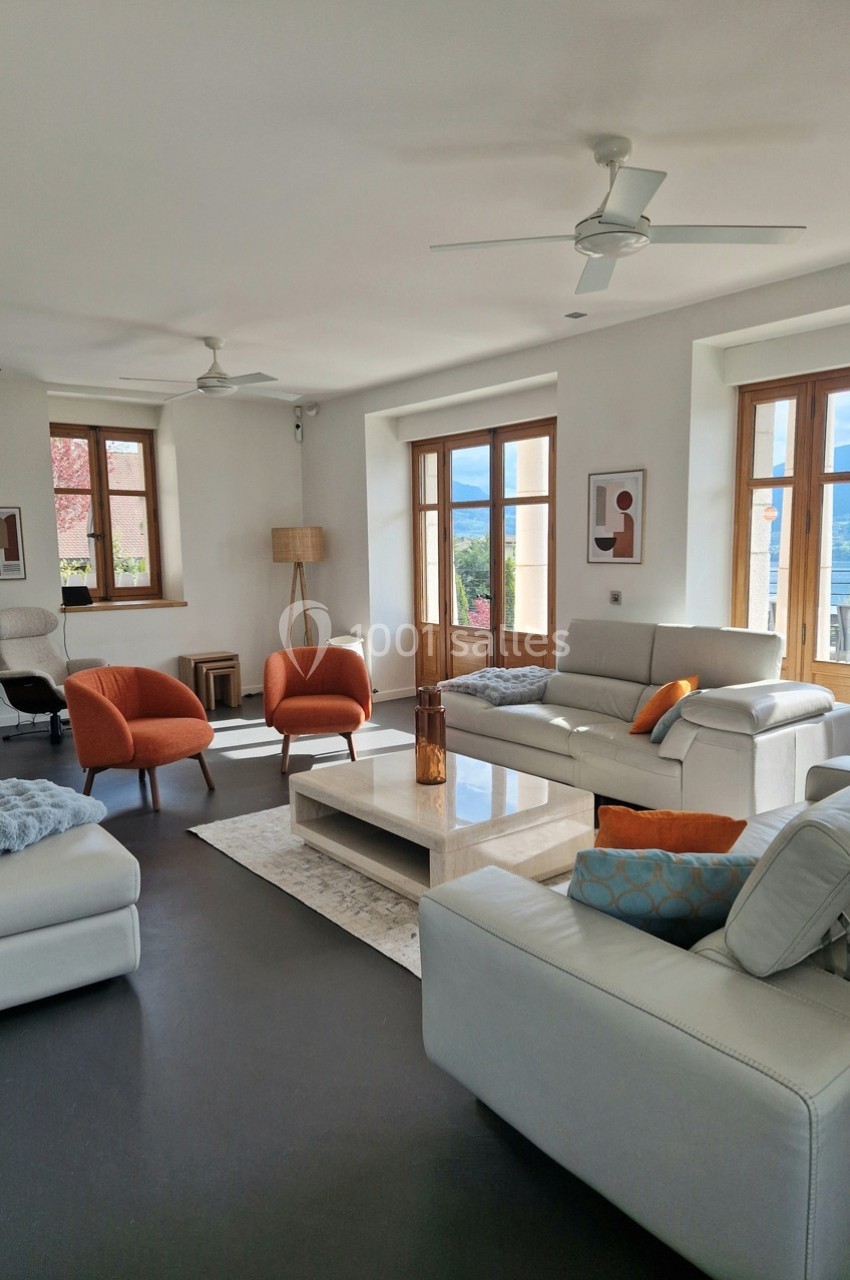 Salon lumineux avec canapés blancs, fauteuils orange, table basse, grandes fenêtres et vue sur un paysage extérieur.