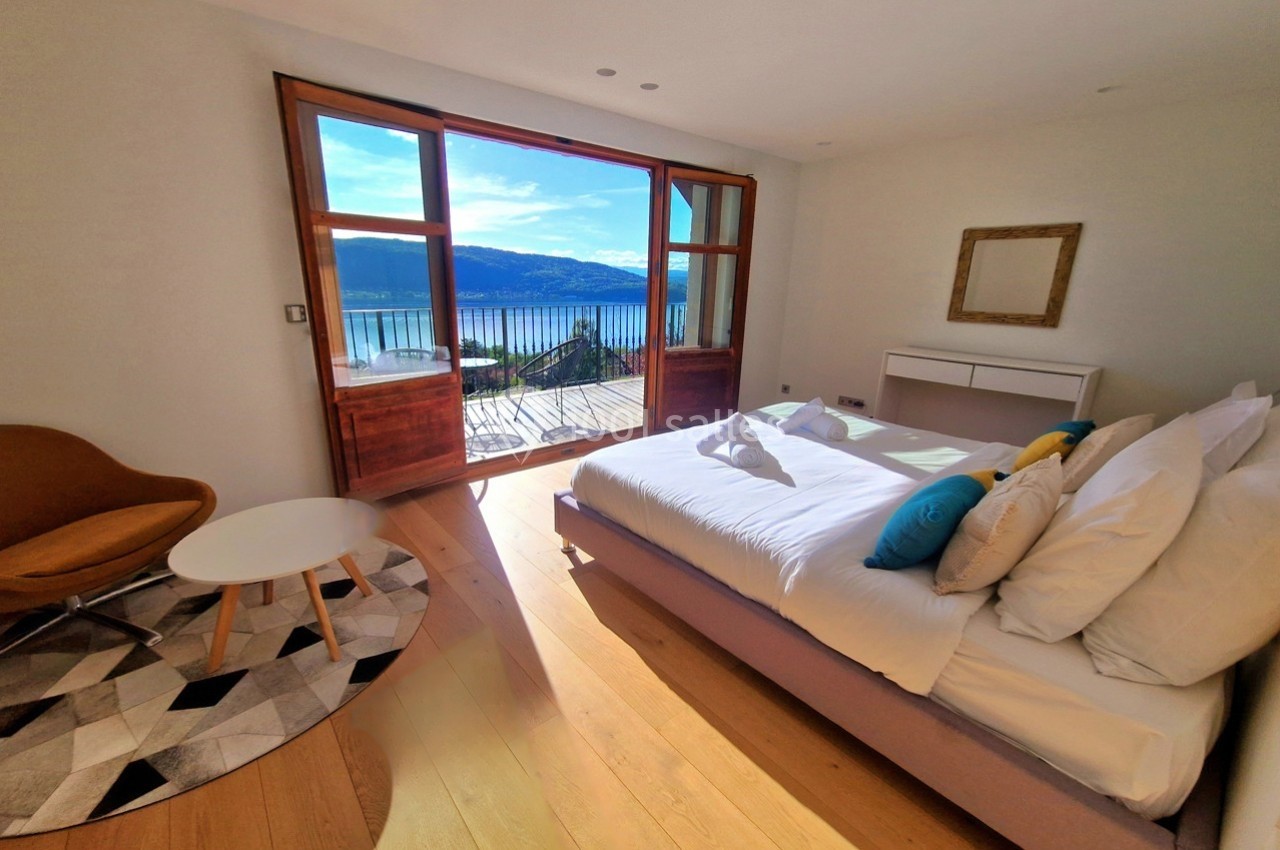 Chambre lumineuse avec lit double, fauteuil, table basse et vue sur un lac à travers une grande baie vitrée.