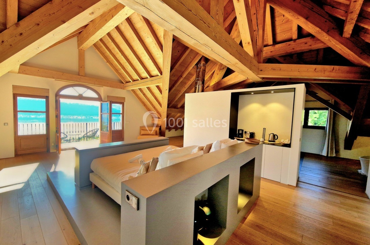 Salon lumineux sous charpente en bois avec canapé, kitchenette moderne et vue sur un balcon donnant sur un paysage.