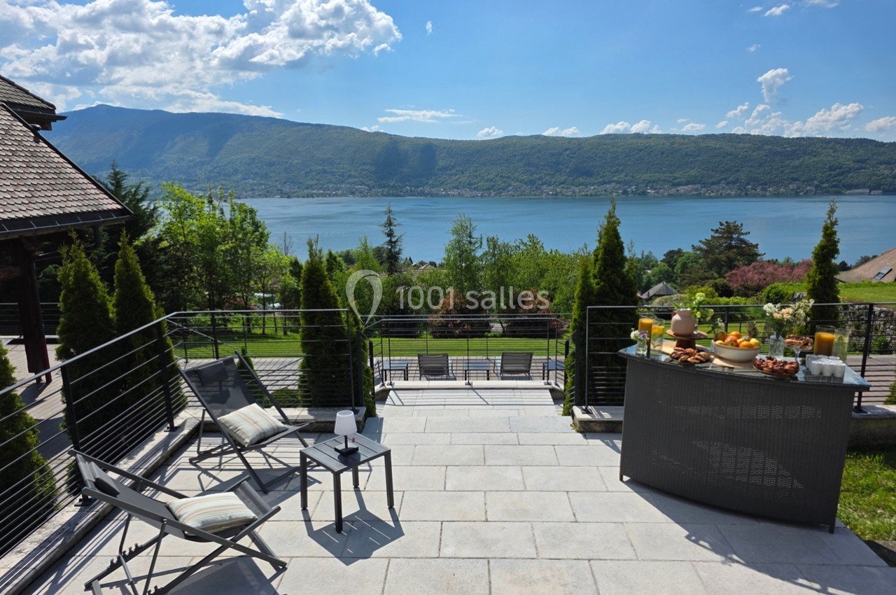 Terrasse avec chaises longues, table garnie de boissons et fruits, vue sur un lac entouré de montagnes et verdure.