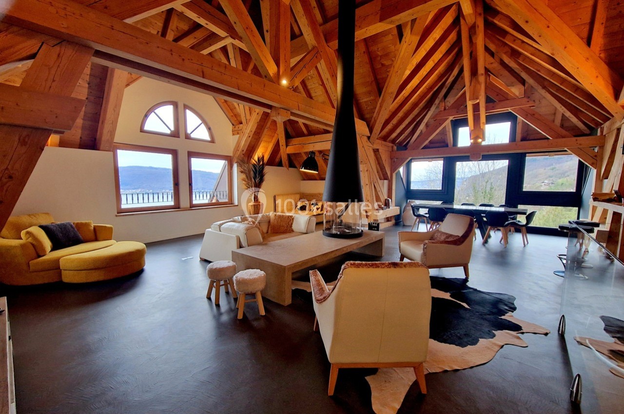 Salon spacieux avec charpente en bois apparente, cheminée centrale, mobilier moderne et grandes fenêtres offrant une vue sur…