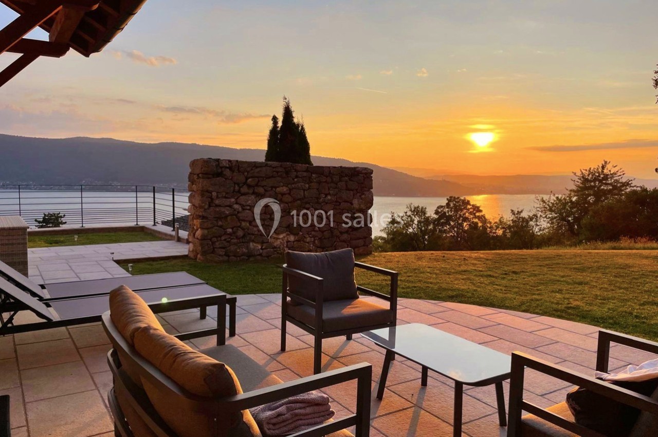 Terrasse avec mobilier de jardin, vue sur un lac au coucher du soleil, entourée de verdure et d'un mur en pierre.