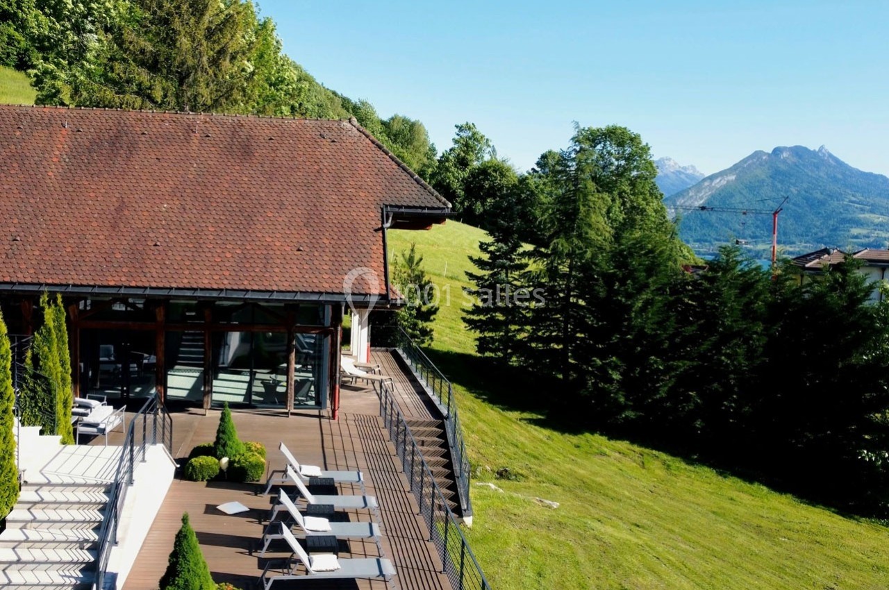 Terrasse en bois avec chaises longues, bordée d'arbres, donnant sur des collines verdoyantes et des montagnes au loin.