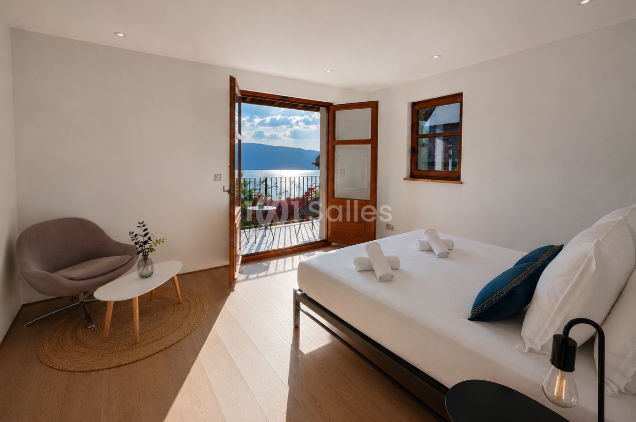 Chambre avec lit blanc, fauteuil taupe et baies vitrées ouvertes sur terrasse avec vue lac et montagne.