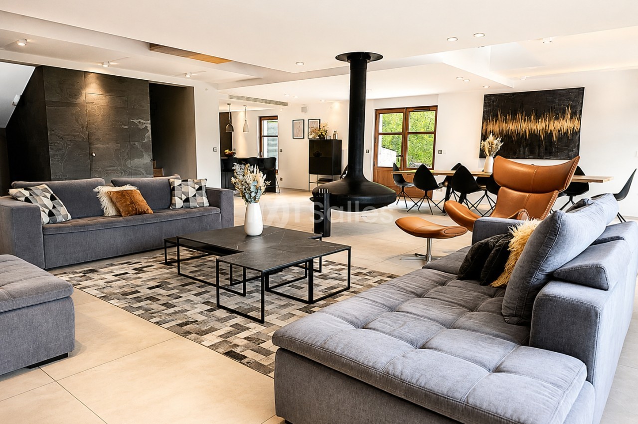Spacieux salon intérieur avec canapés gris, fauteuil cognac, foyer central et baies vitrées donnant sur verdure.
