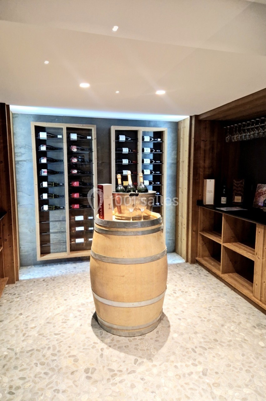 Cave à vin avec étagères remplies de bouteilles, tonneau central servant de table avec verres et bouteilles.