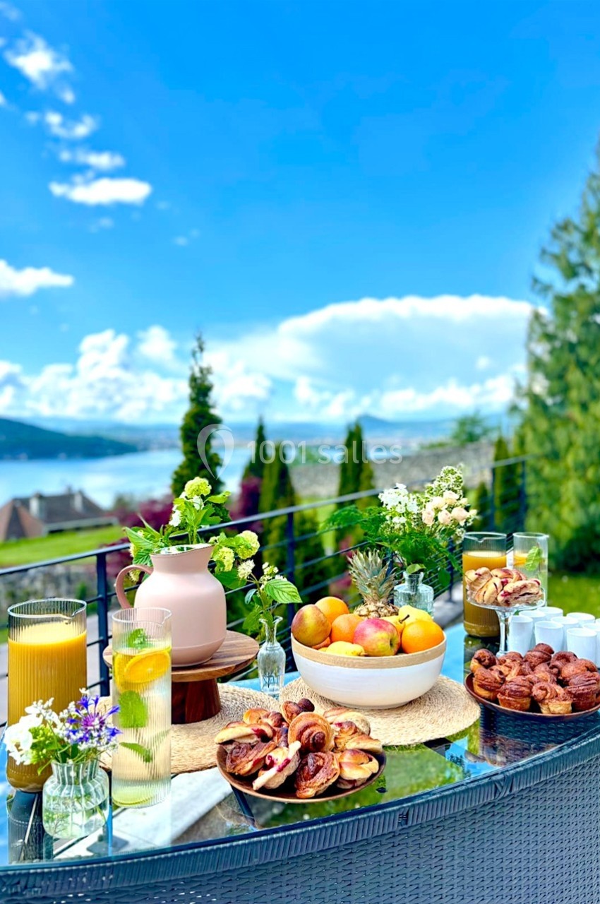 Table extérieure dressée avec fruits, viennoiseries, fleurs et boissons, vue sur un lac et des montagnes en arrière-plan.