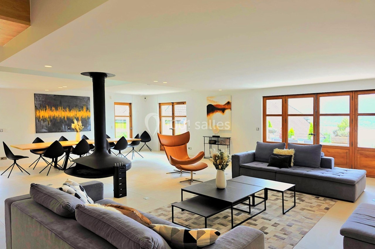 Salon spacieux et lumineux avec canapés, fauteuil design, table à manger, cheminée suspendue et grandes fenêtres.