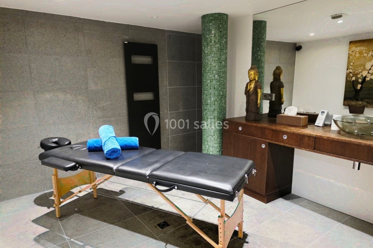 Salle de massage avec table noire, serviettes bleues pliées, statues décoratives et lavabo moderne en arrière-plan.