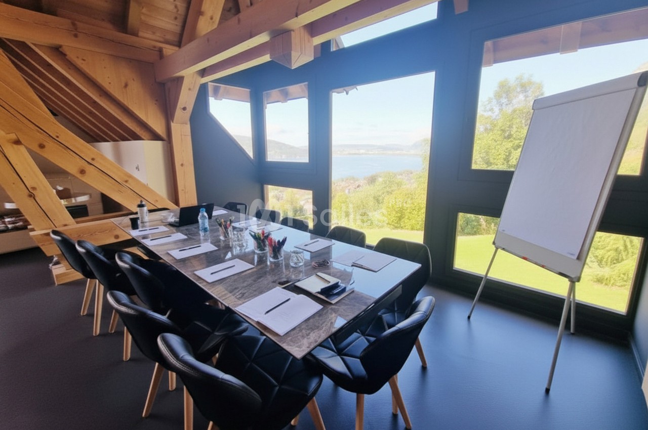 Salle de réunion lumineuse avec table équipée de fournitures, chaises noires et vue sur un paysage verdoyant et un lac.