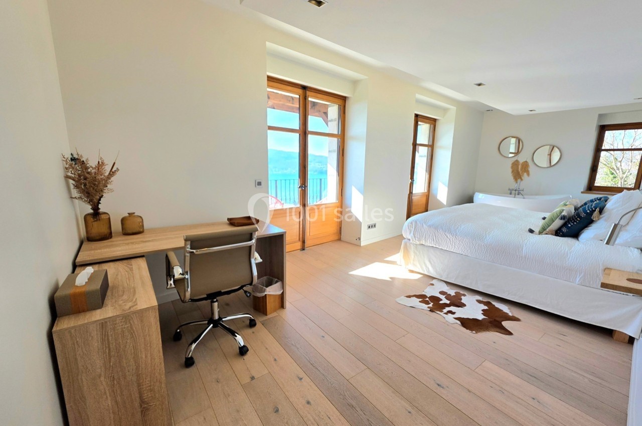 Chambre lumineuse avec lit double, bureau en bois, parquet clair et vue sur un balcon donnant sur un paysage extérieur.