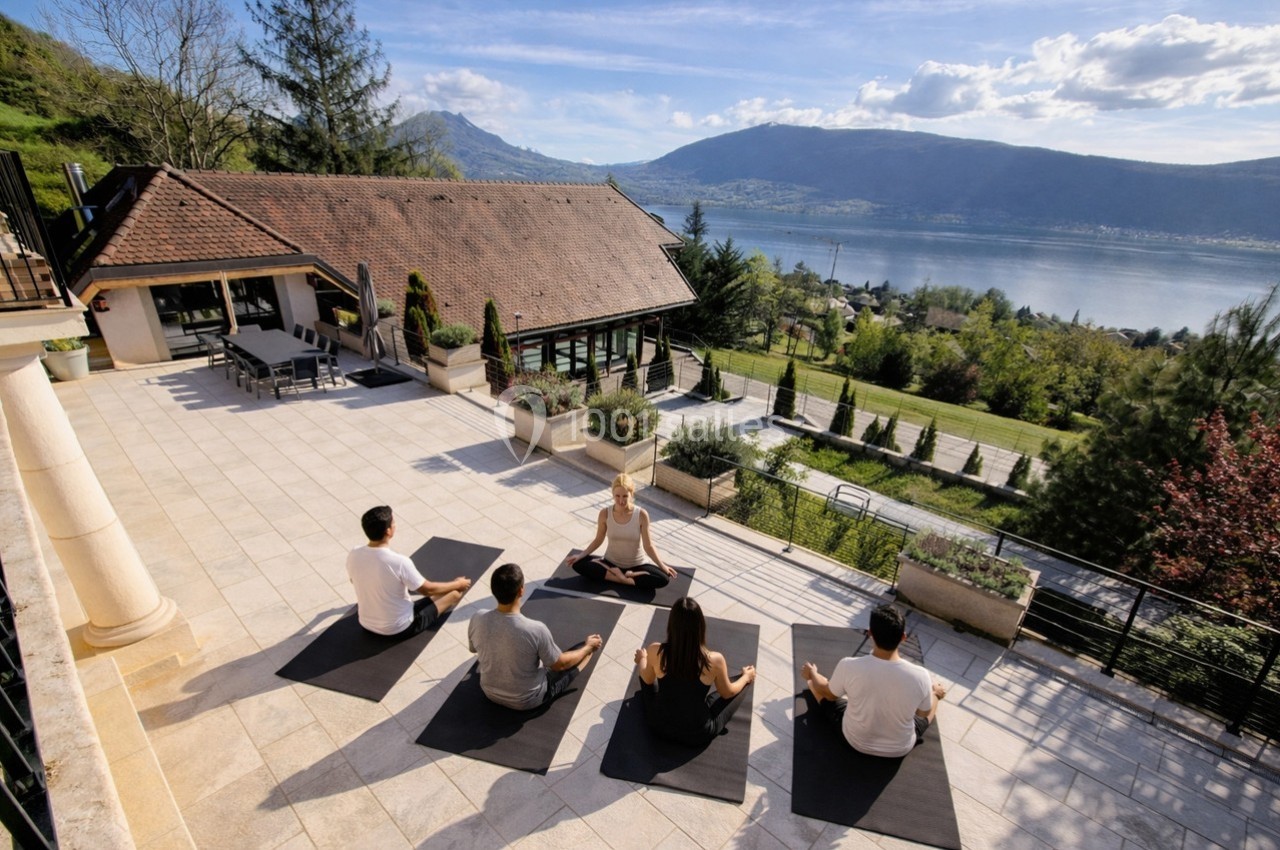 Un groupe de personnes pratique le yoga en plein air sur une terrasse avec vue sur un lac et des montagnes.